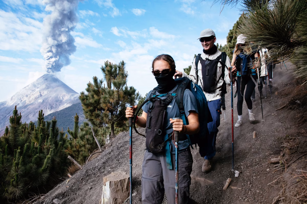 Hikers with face masks walking on volcanic trail with erupting Fuego volcano in background