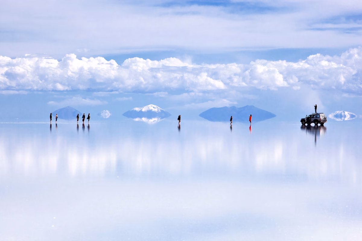 People and vehicle walking on reflective Salar de Uyuni salt flats with mountains mirrored in water